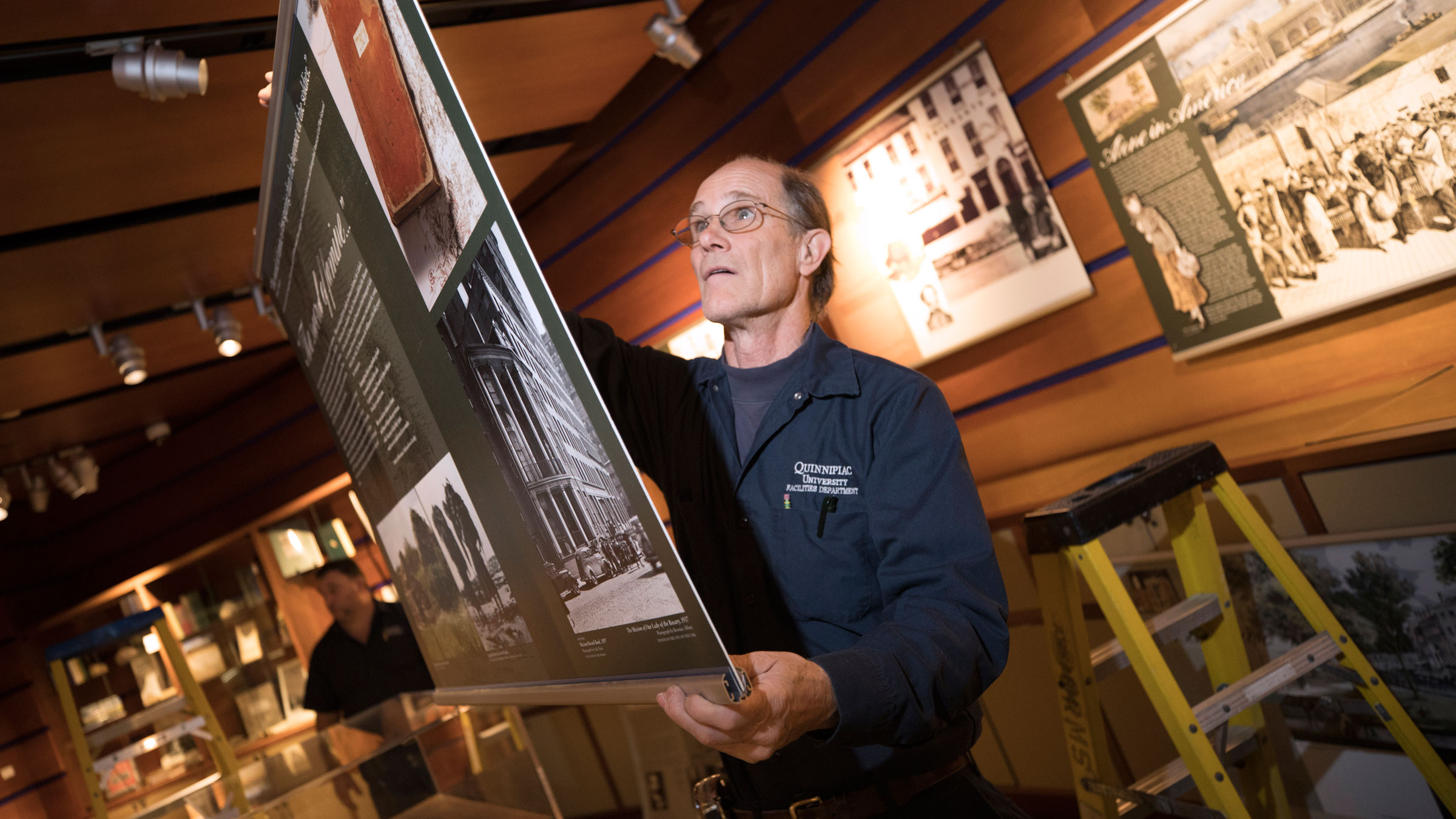 A facilities employee holds up an art piece as he hangs it in advance of an exhibition in the Arnold Bernhard Library.